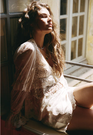 A woman with long wavy brown hair sits by a sunlit window wearing a sheer cream lace robe and matching lace lingerie. The warm sunlight creates a soft, dreamy look with strong shadows across her face and body.