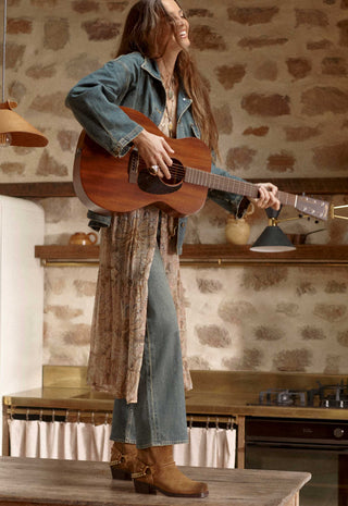 A woman stands in a rustic kitchen setting, holding and playing an acoustic guitar. She is wearing a blue denim jacket, blue jeans, and brown leather boots.