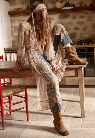 A woman in a bohemian-style patchwork long-sleeve dress and blue jeans sits on a wooden table in a rustic kitchen, wearing brown suede boots.
