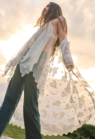 A woman outdoors wearing a sheer white lace long-sleeve cardigan with gold butterfly and moon motifs over dark teal wide-leg jeans, photographed against a bright sunset sky.