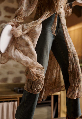 A close-up lifestyle shot of a person wearing a long, flowing beige and brown patterned sheer cardigan or duster over dark blue jeans, holding a white plate in a rustic indoor setting.