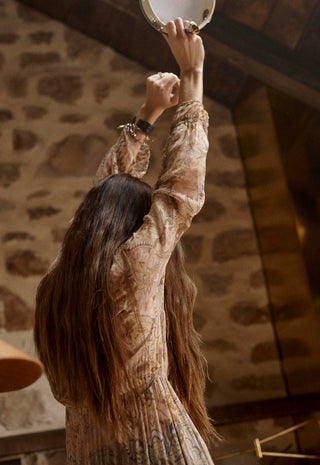 A woman with long dark hair seen from behind, wearing a beige and brown patterned long-sleeve dress, reaching upward in a rustic stone-walled room.