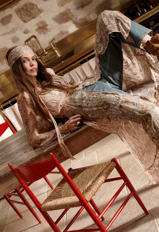 A woman reclines on a wooden table in a rustic interior, wearing a long-sleeve bohemian-style patterned dress in beige, brown, and blue tones with blue jeans. Red chairs are visible in the foreground.