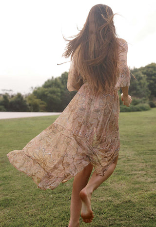 A woman with long brown hair runs barefoot across a grassy field, viewed from behind, wearing a flowing beige bohemian lace dress with a subtle floral pattern.