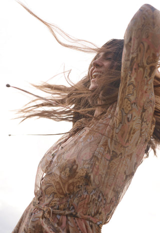 A woman with long, wind-blown brown hair wearing a loose, patterned beige dress with brown and pink paisley-like motifs, captured against a bright white sky.