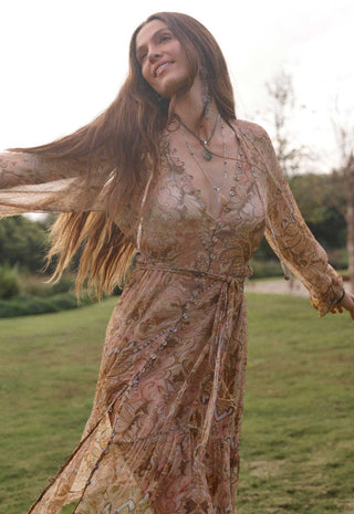 A woman outdoors in a grassy park wearing a flowing sheer boho-style maxi dress with an intricate paisley and floral pattern in warm beige, brown, and muted pink tones. She is captured mid-spin with her hair blowing, wearing layered necklaces.
