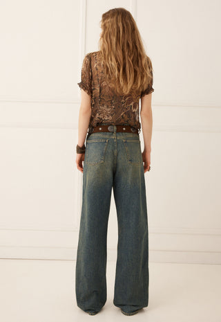 A woman viewed from behind wearing a brown patterned short-sleeve top, a brown belt, and wide-leg blue jeans against a white paneled wall.