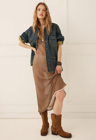 A woman models a brown midi slip dress with a sheer overlay, styled with a blue denim shirt jacket, brown leather boots, and a brown choker against a white paneled wall.
