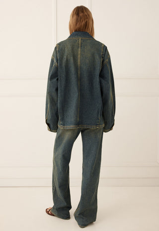 A model viewed from the back wearing an oversized dark blue denim jacket and matching wide-leg denim jeans, standing against a white paneled wall.