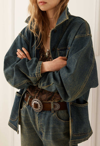 A person wearing an oversized dark blue denim jacket with wide sleeves, paired with a brown patterned top, a brown leather belt with a large silver concho buckle, and blue jeans, set against a white paneled background.
