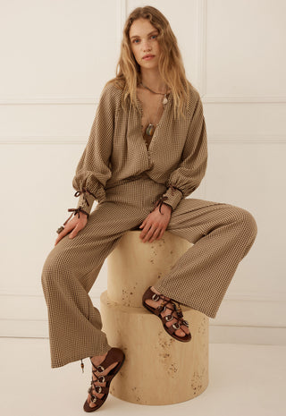 A woman sits on a round stone pedestal wearing a brown and beige gingham two-piece set consisting of a long-sleeve blouse and wide-leg trousers