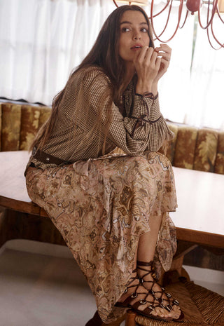 A woman sits on a wooden bench in a warm, sunlit room, wearing a sheer patterned brown and beige blouse with a matching long skirt, lace-up strappy sandals, and bracelets.