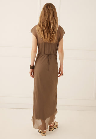 A woman viewed from behind wearing a brown midi dress with short cap sleeves, a gathered waist, and a drawstring tie, paired with platform sandals against a white paneled wall.