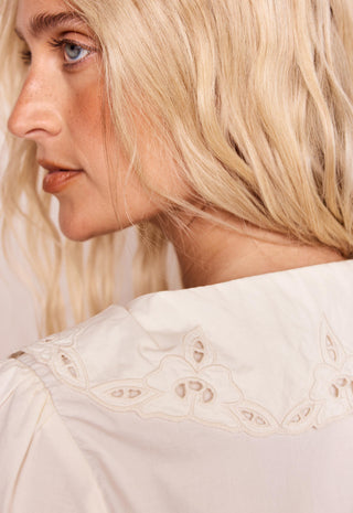 Close-up side profile of a blonde woman wearing a cream-colored blouse with delicate eyelet embroidery around the collar.