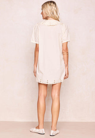 A woman shown from the back wearing a short-sleeve cream-colored tunic dress with a ruffled collar and eyelet lace trim at the hem, paired with white lace-up flats, against a textured beige wall.