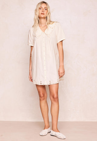 A woman modeling a short-sleeve cream-colored mini dress with a ruffled Peter Pan collar and eyelet lace trim, paired with white lace-up flats, against a textured beige wall.