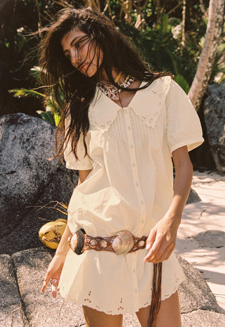 A woman outdoors wearing a cream-colored short-sleeve button-front dress with eyelet trim and a matching belt, standing among rocks and tropical plants.