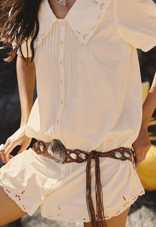 A close-up of a person wearing a cream short-sleeve blouse with eyelet lace trim and matching cream shorts, styled with a brown woven belt featuring cowhide and shell details.