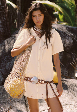A woman stands outdoors on rocky ground wearing a cream short-sleeve button-front dress or tunic with a fringed hem