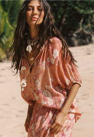 A woman on a sandy beach wearing a loose, dusty pink floral dress with voluminous short sleeves and a draped neckline.