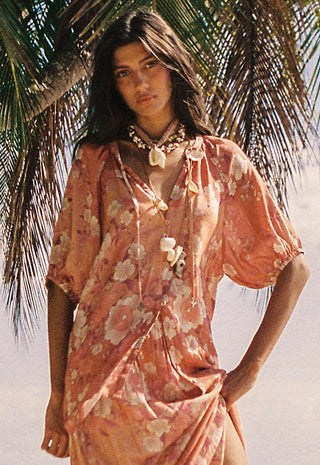 A woman wearing a loose, peach floral cover-up dress with shell-like tie details, standing outdoors near palm leaves against a bright beach-like background.