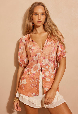A woman modeling a short-sleeve button-up blouse with a peach, pink, and white floral print, paired with white ruffled shorts, against a warm beige background.