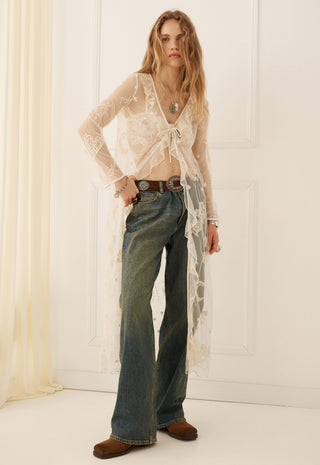 A woman stands indoors wearing a sheer white lace long-sleeve cardigan over a cropped top
