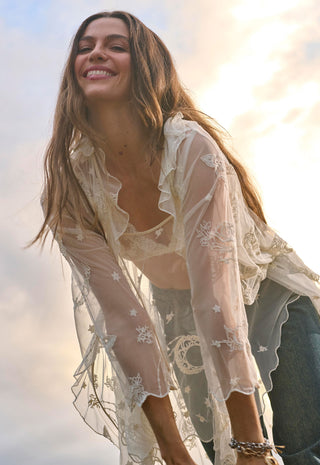 A woman outdoors wearing a sheer cream lace bohemian-style cover-up over a matching bralette and jeans, with a bright sky background.