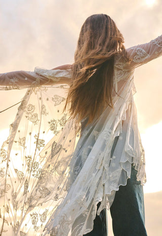 A woman seen from behind wearing a sheer white lace long-sleeve duster or wrap with butterfly and floral embroidery, paired with dark jeans, set against a bright, sunlit sky.