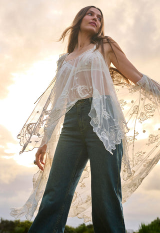 A woman wearing a sheer white floral lace long-sleeve top over a white camisole and dark blue wide-leg jeans, photographed outdoors against a bright sky with the sun shining.