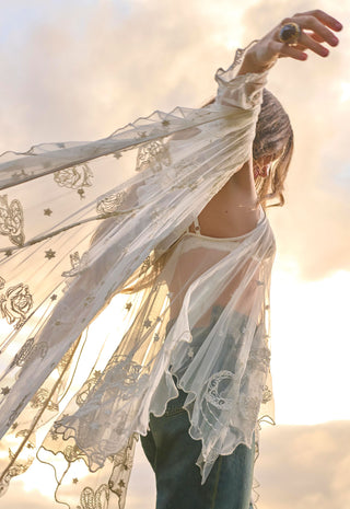 A woman outdoors wearing a sheer white embroidered lace flutter-sleeve cardigan over a white top and blue jeans, with her arm raised against a bright sunset sky.
