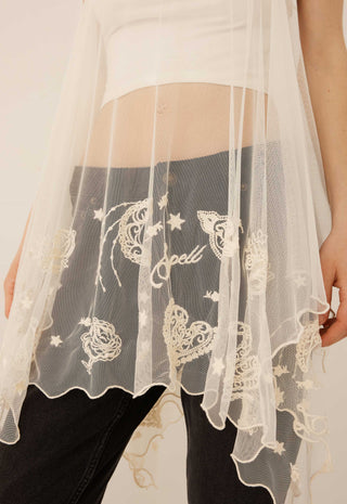 A close-up of a person wearing a sheer white lace overlay draped over dark gray jeans, with a white crop top partially visible. The lace features embroidered celestial motifs including stars, hearts, and the word 'spell' in script.