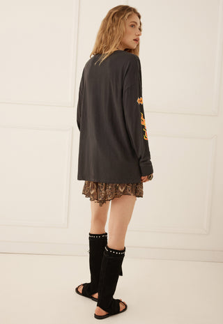 A woman is shown from the back wearing an oversized black long-sleeve top with orange graphic print on the sleeve, a brown patterned mini skirt, and black knee-high studded boots against a white paneled wall.