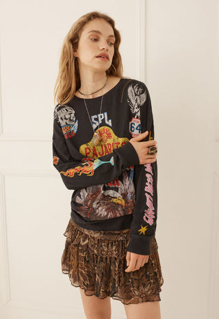 A woman modeling a black long-sleeve graphic sweatshirt with colorful vintage-style prints and a brown patterned ruffled mini skirt, standing against a white paneled wall.